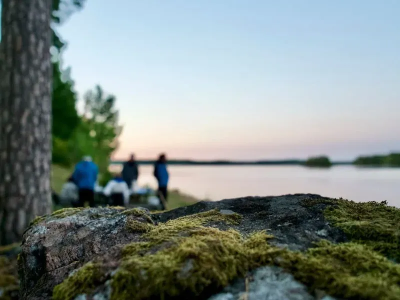 Mossig sten vid en sjö i kvällsljus, med människor vid stranden